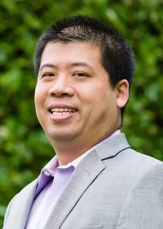 Orthopedist Dr. Clifford Mah smiling in a gray suit and tie in front of green shrubbery